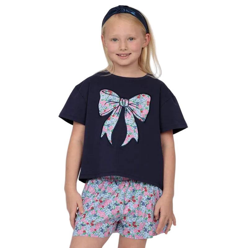 Young girl wearing a navy blue t-shirt with a large pink bow design and floral shorts on a white background.