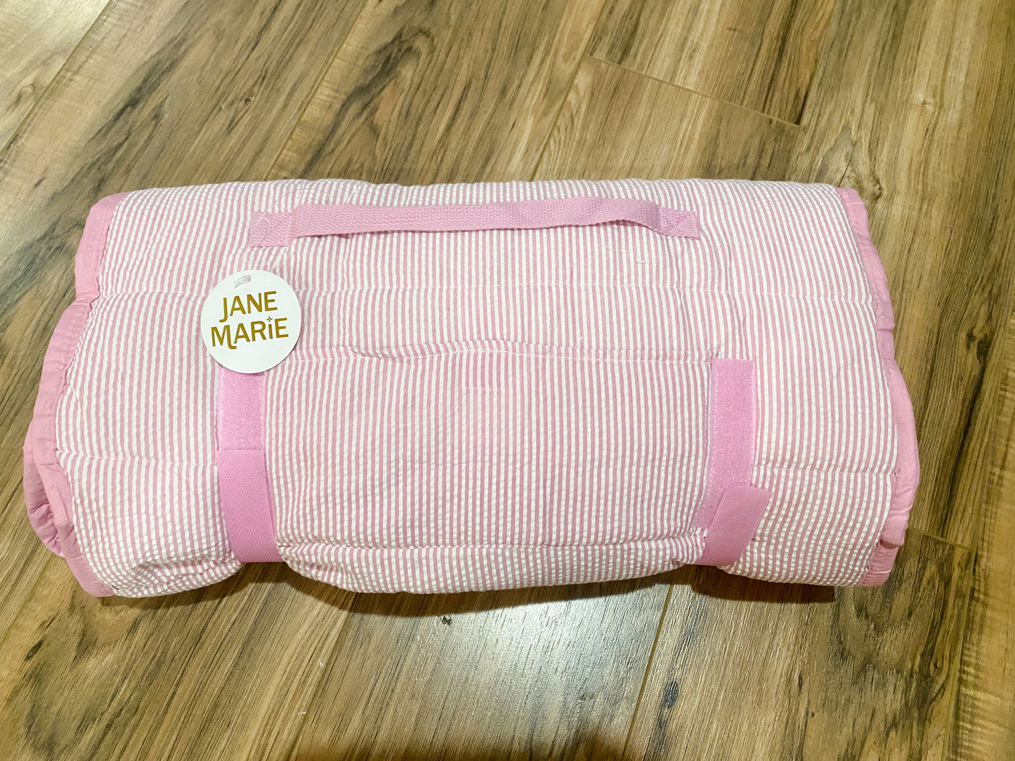 A rolled-up quilted blanket with pink and white stripes and solid pink trim, featuring a circular white tag that reads 'JANE MARIE' in gold lettering, placed on a wooden floor.