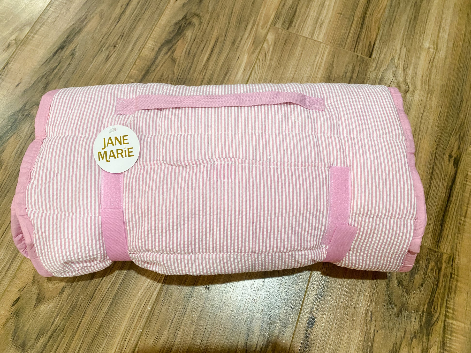 A rolled-up quilted blanket with pink and white stripes and solid pink trim, featuring a circular white tag that reads 'JANE MARIE' in gold lettering, placed on a wooden floor.