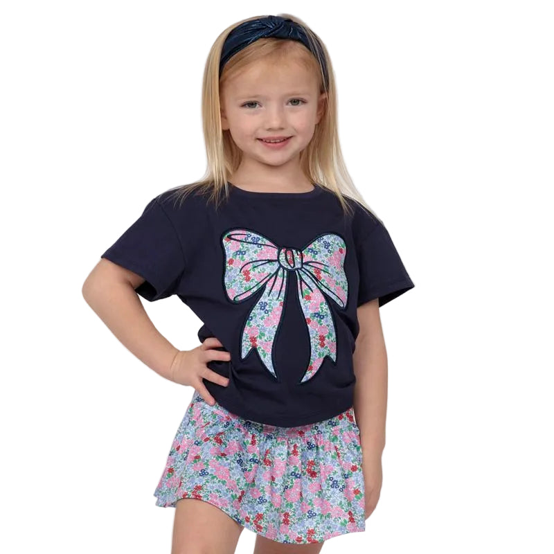 Young girl wearing a navy blue t-shirt with a floral bow design and a matching skirt on a white background.